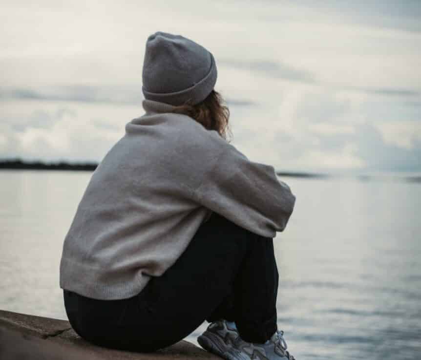 A girl sitting on a rock by the water in a grey hoodie and toque, hosted by Wellin5 in Canada.