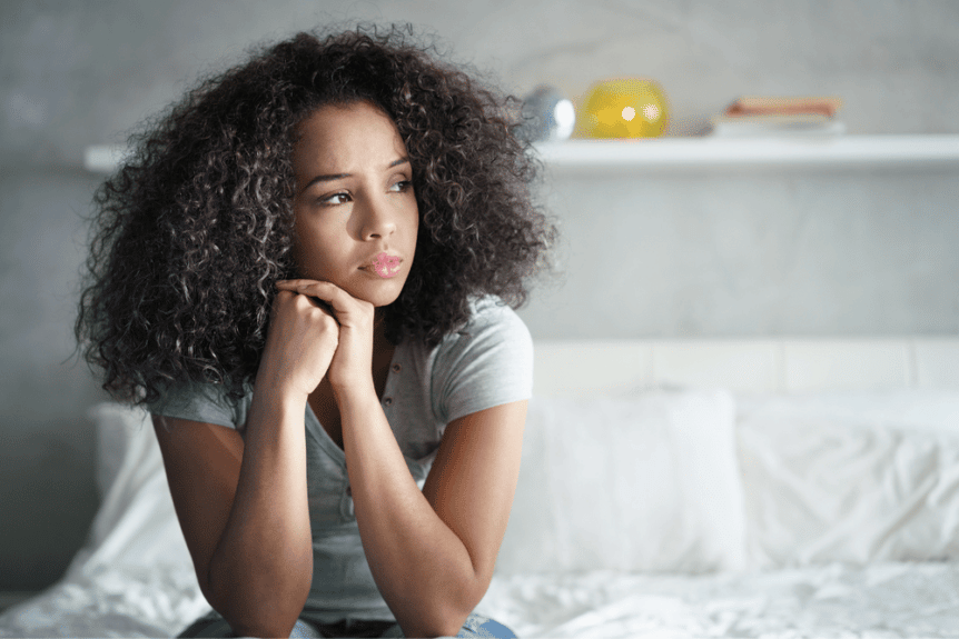 A young woman sits on her bed, looking away from the camera with a sad expression. 3 signs of depression in women hosted by Wellin5.