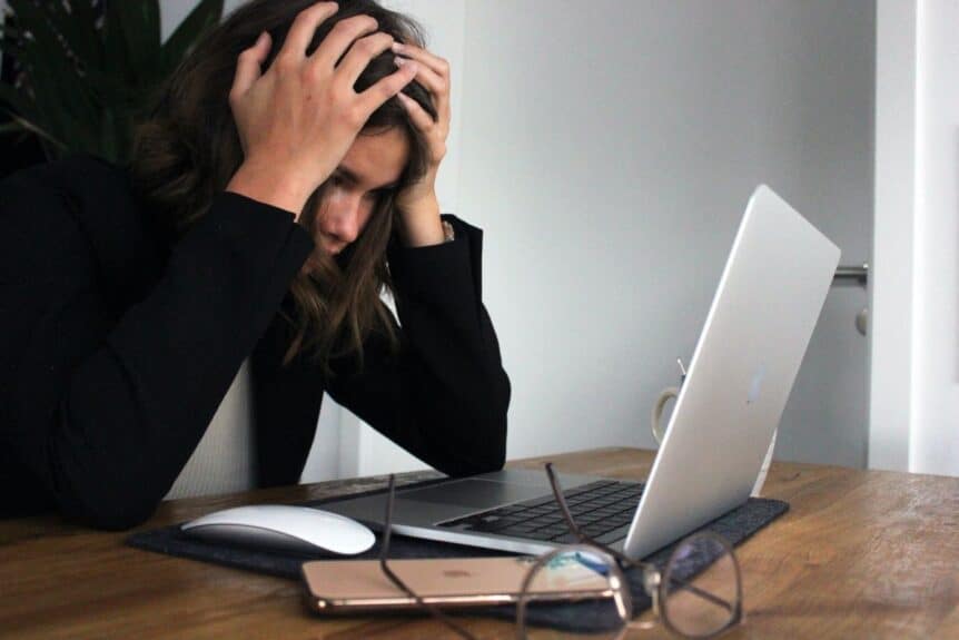 A woman sitting in front of her laptop with head in hands, looking stressed. 7 of the most common anxiety triggers hosted by Wellin5 online counsellors across canada.